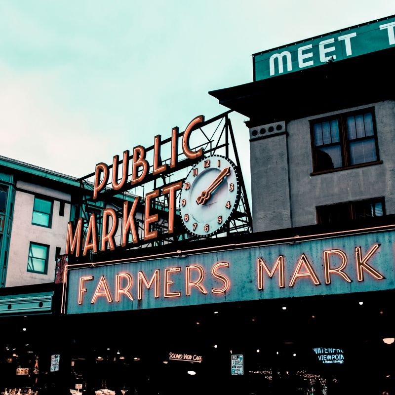 The image shows the iconic "Public Market" and "Farmers Market" signs with a clock, situated between two buildings in an urban area under a cloudy sky.