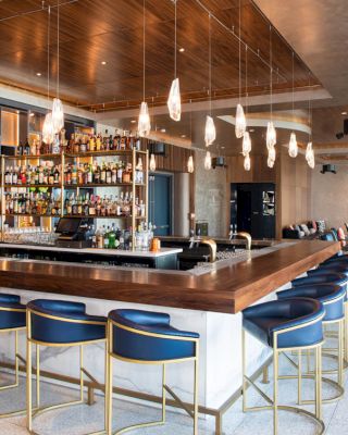 A modern bar with a wooden countertop, blue bar stools, hanging lights, and a fully stocked back bar, located in a stylish, open lounge area.