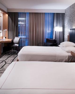 A modern hotel room with two double beds, a wall-mounted TV, a desk with a chair, and ambient lighting.