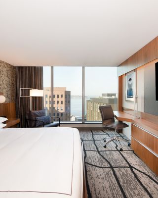 This image shows a modern hotel room with a large bed, a desk, a chair, a TV on the wall, and a window offering a cityscape view.