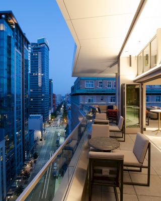 The image shows a modern balcony with elegant furniture, overlooking a city skyline of tall buildings at dusk. The scene is well-lit.