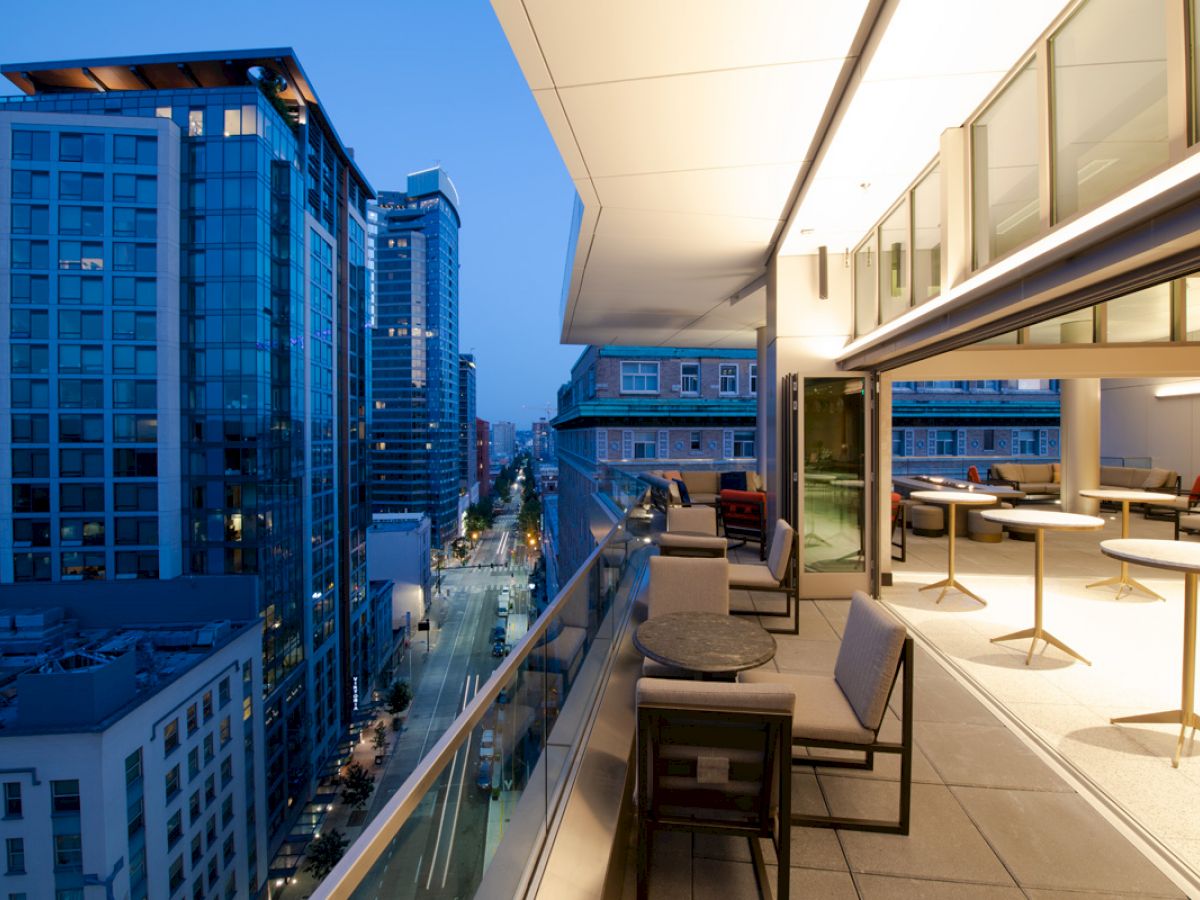 A modern cityscape at dusk features a high-rise balcony with outdoor seating and tables, overlooking a busy urban street lined with buildings.
