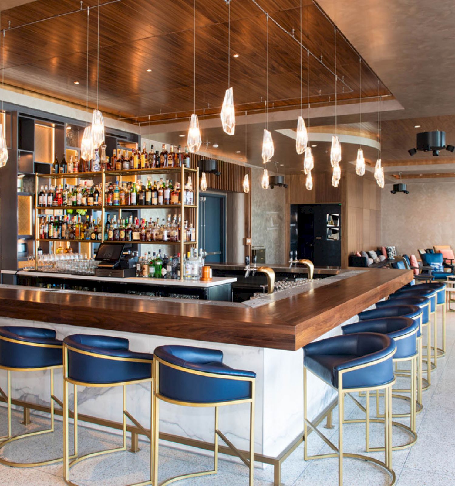 A modern bar with blue stools, contemporary lighting, and a well-stocked shelf of bottles. The space features a lounge area in the background.