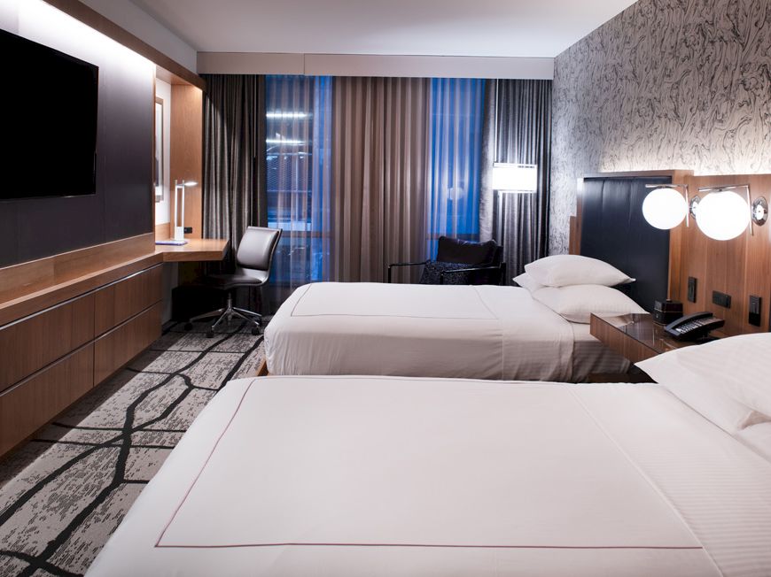 A modern hotel room with two beds, a wall-mounted TV, a desk, chairs, and lamps on bedside tables and on the desk.