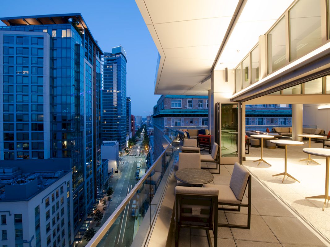 A modern cityscape viewed from a balcony at dusk, with tall buildings and a furnished outdoor seating area on the right side of the image.