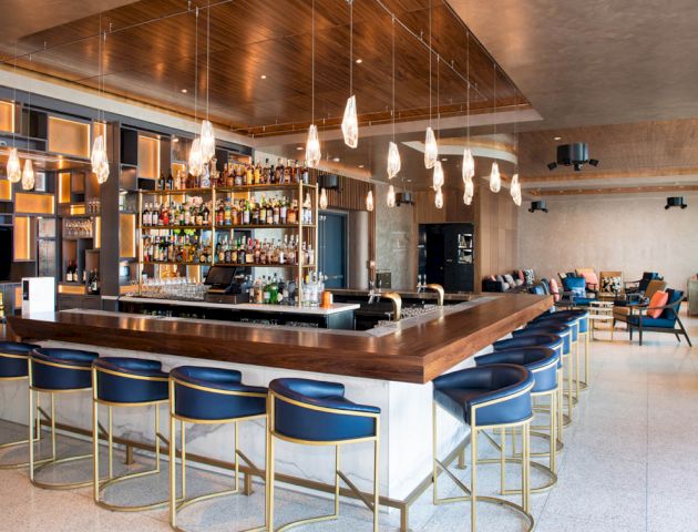 The image shows a modern bar with blue cushioned stools, sleek wooden counters, hanging lights, and a well-stocked liquor display in a cozy setting.