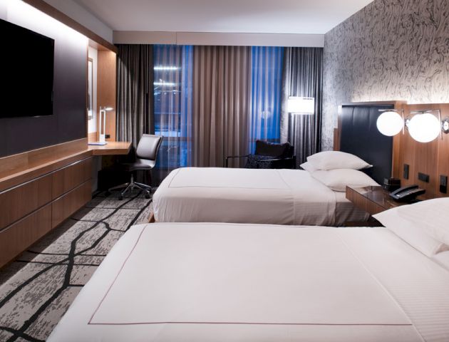 A modern hotel room features two neatly made beds, a flat-screen TV, a desk with a chair, contemporary lighting, and large window curtains.