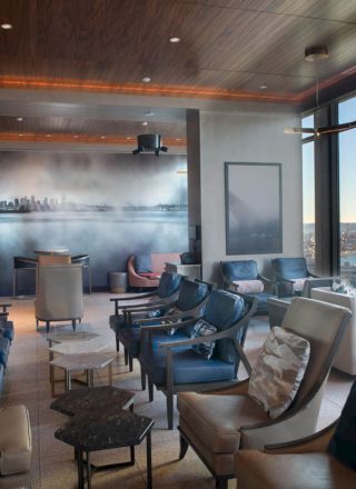 A modern lounge area with large windows offering a coastal view, featuring comfortable seating, a mural on the back wall, and sleek decor.