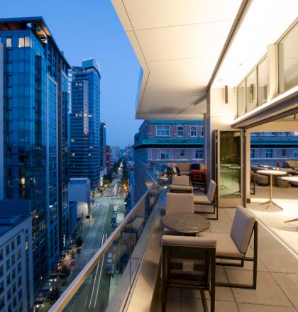 A modern urban balcony at dusk, with seating and tables, overlooks city streets and tall buildings bathed in evening light.