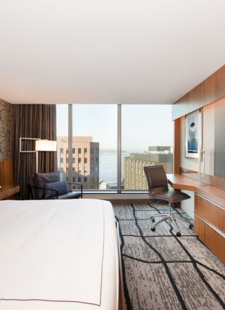 The image shows a modern hotel room with a large bed, wall-mounted TV, desk, and chair. There's a cityscape view through the large window.