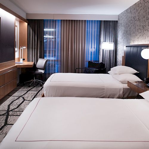A modern hotel room with two beds, a wall-mounted TV, a desk, and a chair against large windows with curtains.