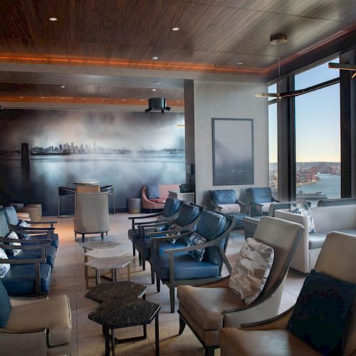 A modern lounge with leather chairs, panoramic windows offering a city and waterfront view, and contemporary wall art.