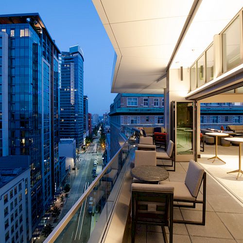 An urban balcony overlooks a cityscape at dusk with modern buildings and a street. The balcony has seating and tables, illuminated by indoor lights.