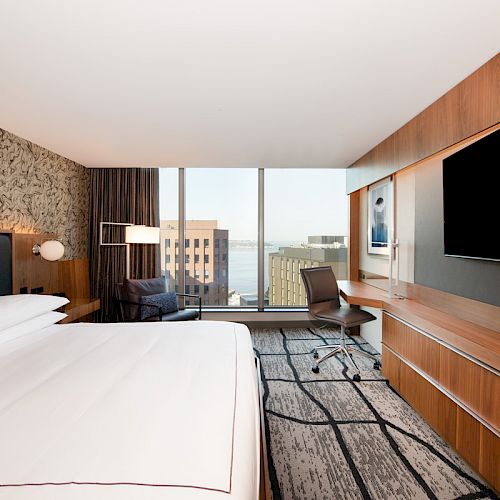 A modern hotel room with a bed, wall-mounted TV, desk, chair, and a large window with a city view in the background.