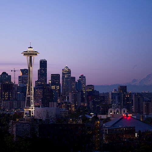 City skyline at dusk featuring the Space Needle, surrounded by high-rise buildings, with a mountain in the background and illuminating city lights.