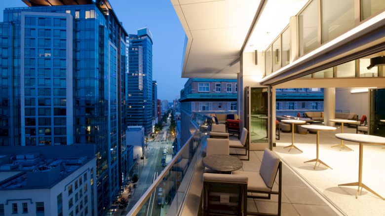 A modern balcony at dusk has a city view with tall buildings. The balcony has tables, chairs, and glass railings.