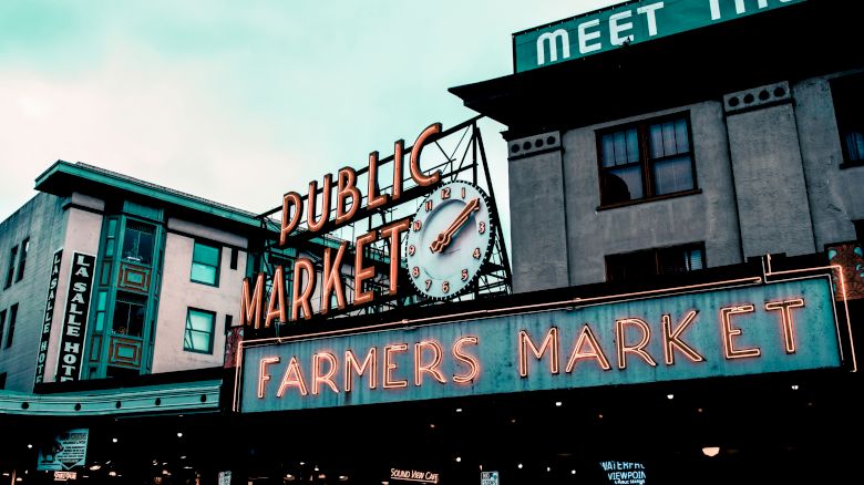 The image shows the exterior of a public market with neon signs reading "Public Market" and "Farmers Market." A clock is also visible on the sign.