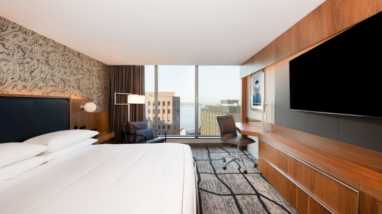 A modern hotel room with a king-sized bed, large TV, desk, and chair, featuring a window with a city view and contemporary decor.
