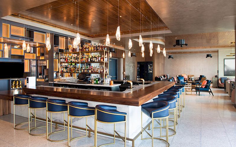 The image shows a modern bar with a well-stocked back shelf, several bar stools, hanging lights, and seating areas in a spacious, contemporary setting.