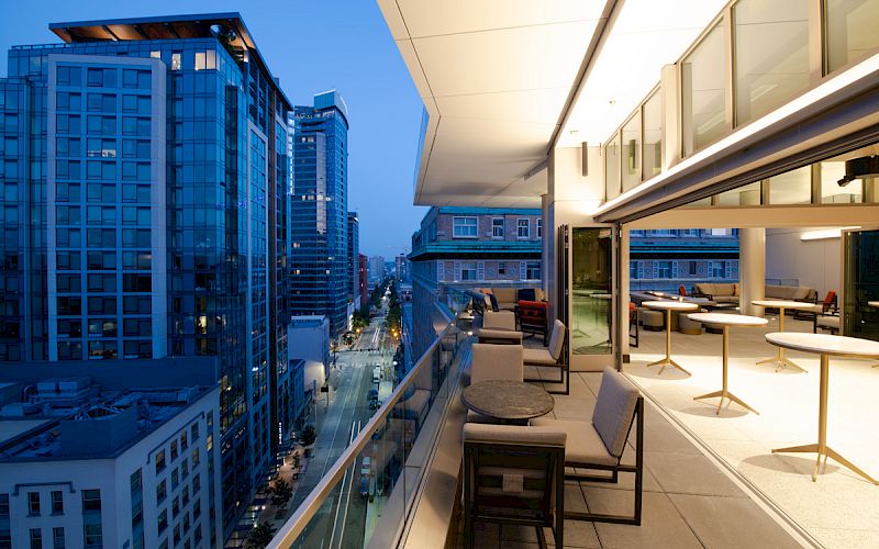A modern balcony with tables and chairs overlooks a cityscape with tall buildings under a twilight sky.