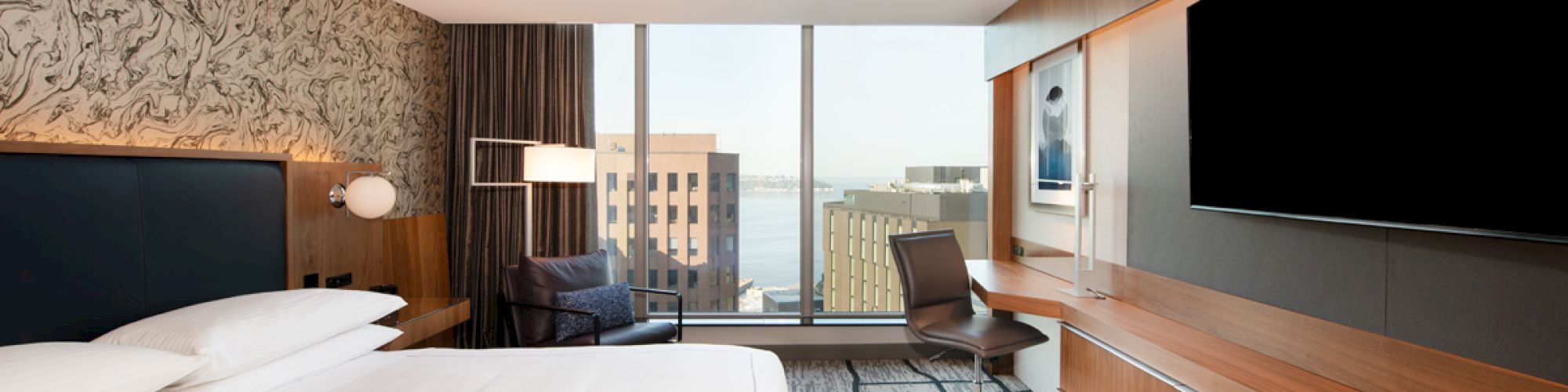 This image shows a modern hotel room with a large bed, a desk and chair, a flat-screen TV, and a window offering a cityscape view.