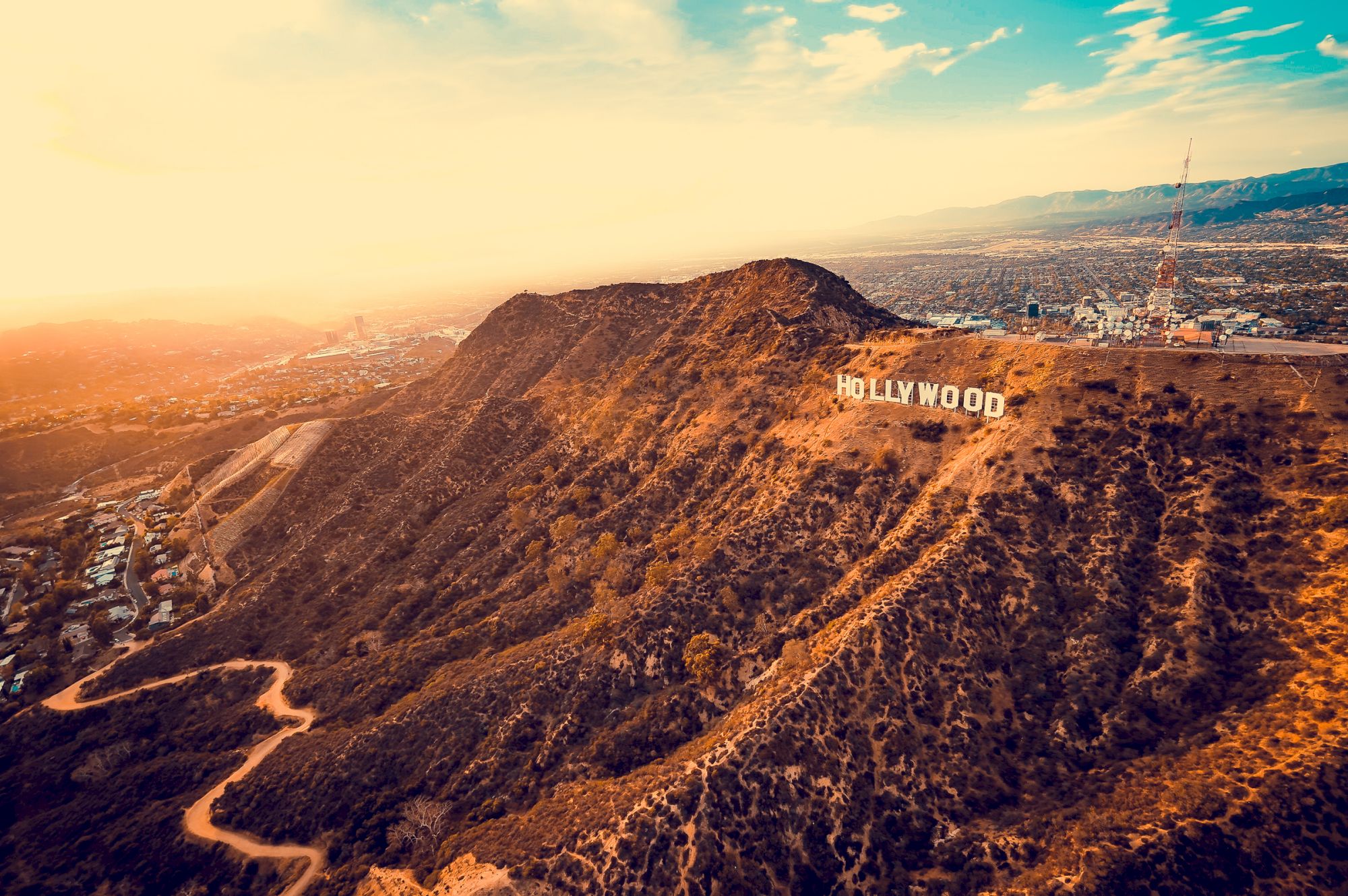 The image shows the iconic Hollywood sign on a hill overlooking Los Angeles, with a scenic landscape of dry hills and a sprawling city below.