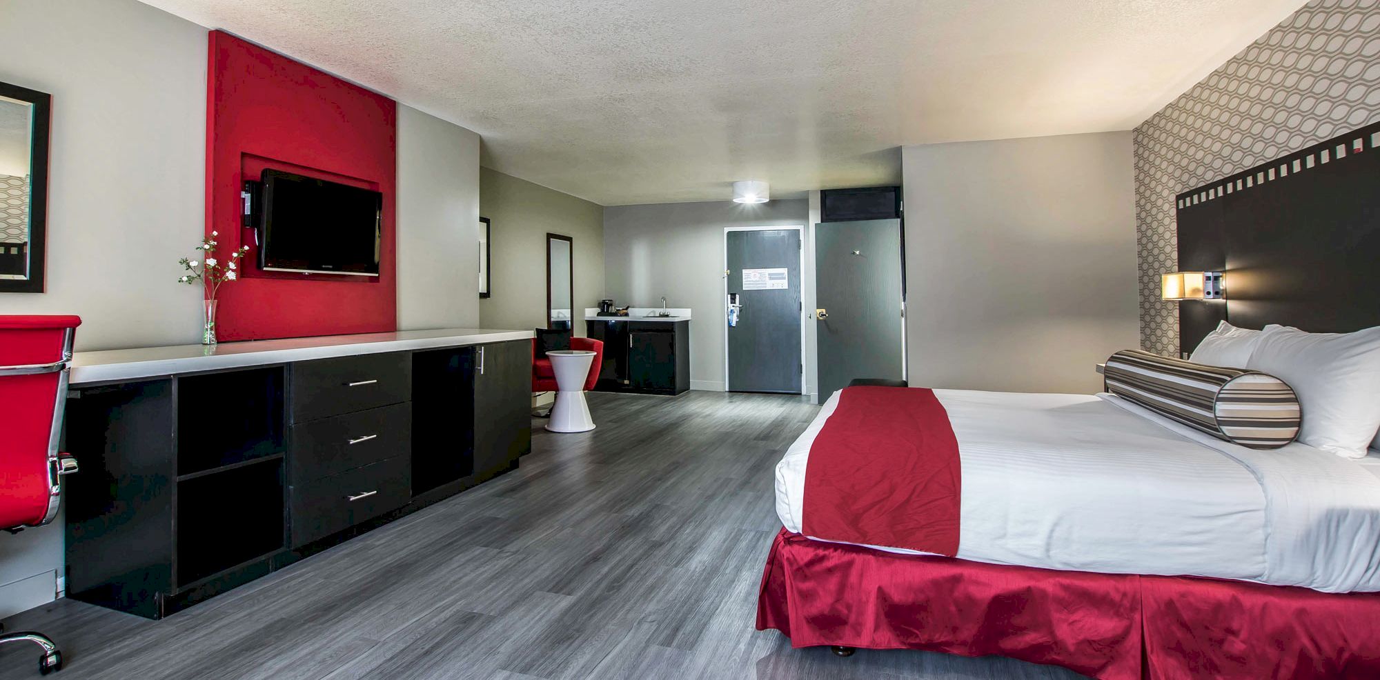 A spacious hotel room with a double bed, red and white decor, a TV, desk, and seating area. Modern design and clean ambiance.