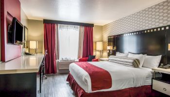 A modern hotel room featuring a large bed with red accents, a work desk, flat-screen TV, armchair, window with curtains, and stylish decor.