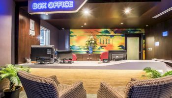 The image depicts a box office with a modern, colorful interior design, including two chairs, a counter, and computer equipment on the desk.