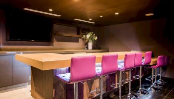 A modern bar setup with a wooden counter, pink bar stools, a flower vase, and overhead lighting creating a cozy ambiance.