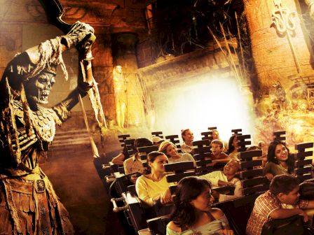 A theme park ride featuring a large skeleton wielding a weapon as passengers look on with mixed emotions in an ancient temple setting.