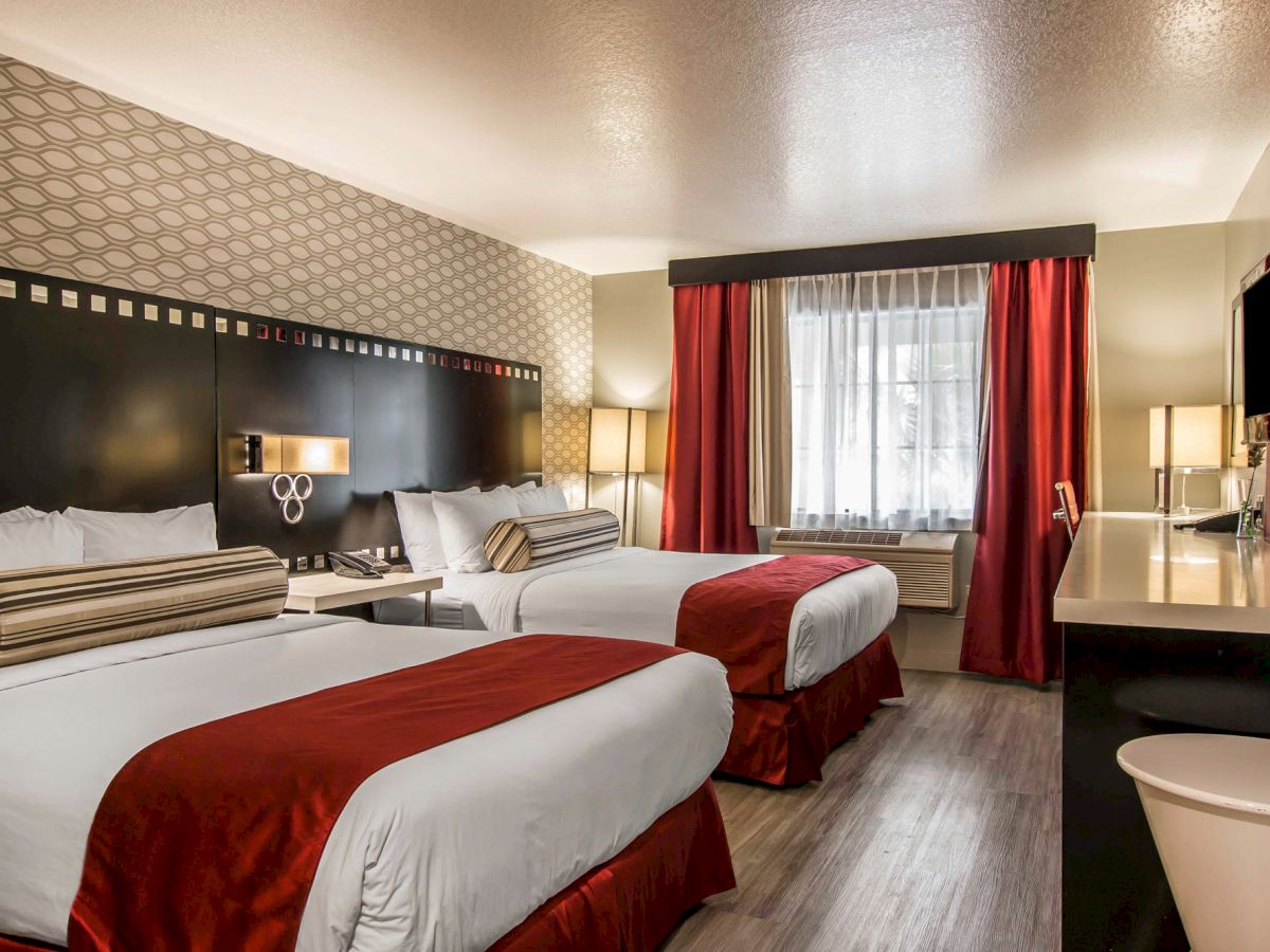 A hotel room with two double beds, featuring red and white bedding, a modern decor, wall-mounted TV, a work desk, and window with red curtains.