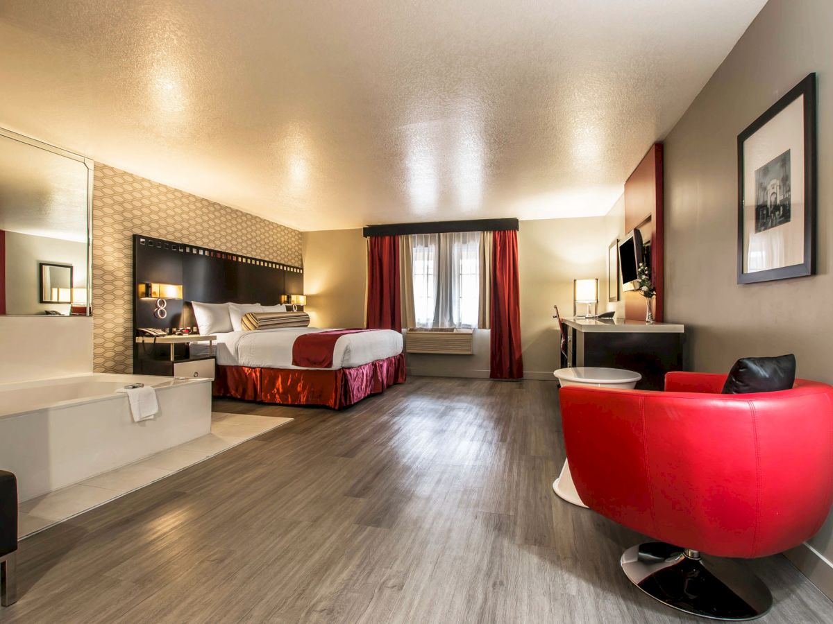 A spacious hotel room with a bed, a red chair, a desk, and modern furnishings. A bathtub is visible next to the bed.