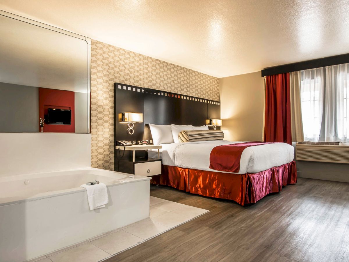 A hotel room featuring a large bed with red and white bedding, a bathtub, a window with curtains, and wooden flooring.